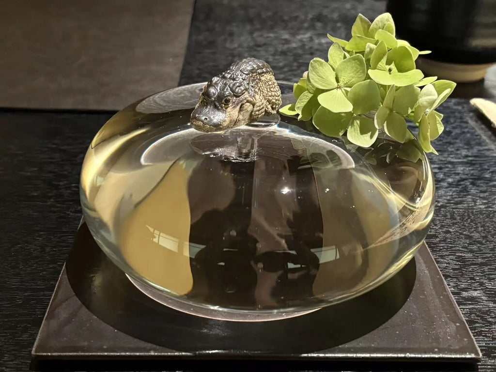 A clear glass dish containing water, with a small sculpted alligator tea pet on top and green hydrangea flowers on the rim, set on a dark square plate against a dark tabletop.