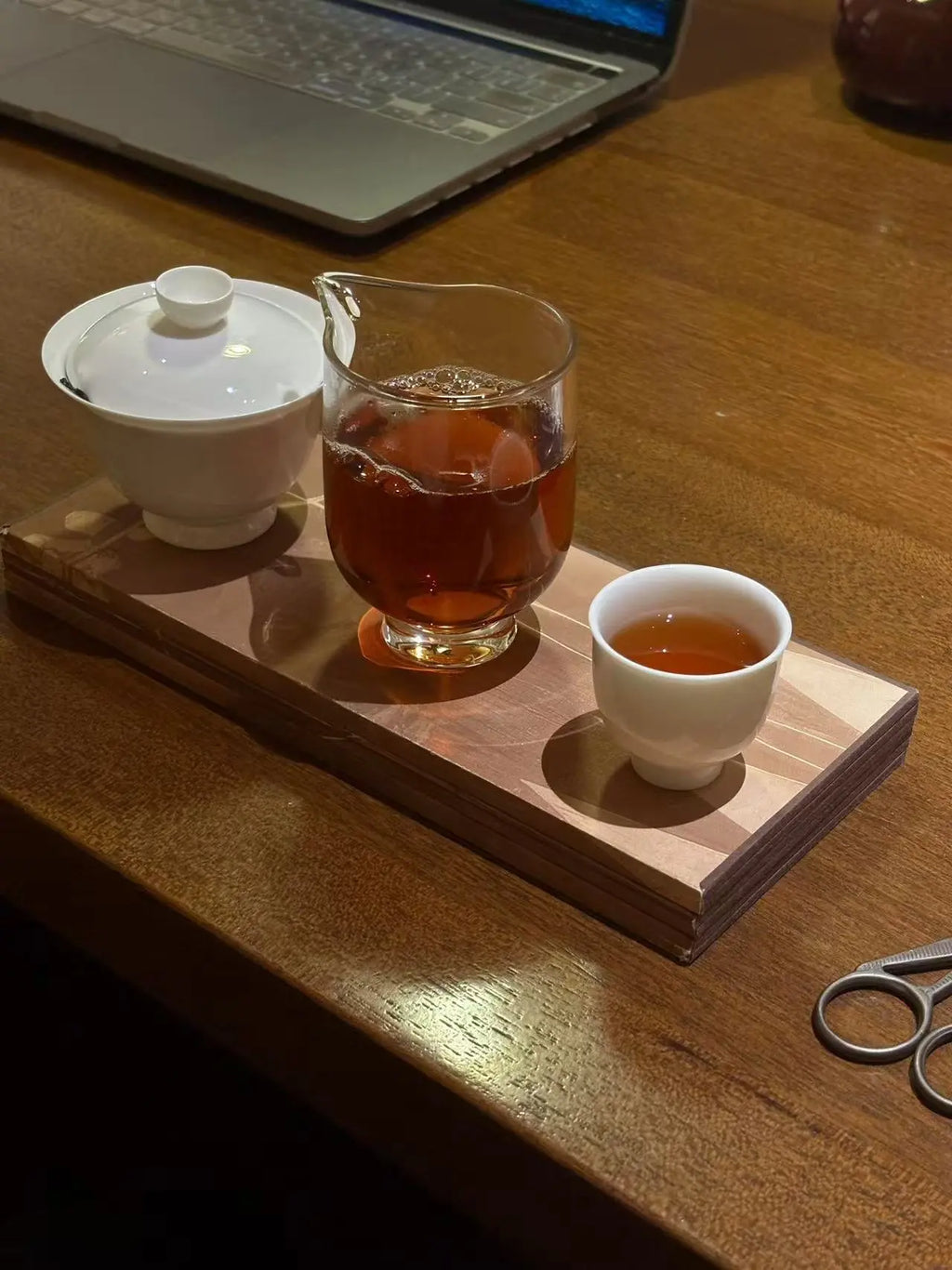 A tea set arrangement on a wooden tray, including a white lidded teapot bowl, a clear glass pitcher filled with amber Cinnamon Rock tea, and a small white cup with tea, placed on a wooden table near a laptop.