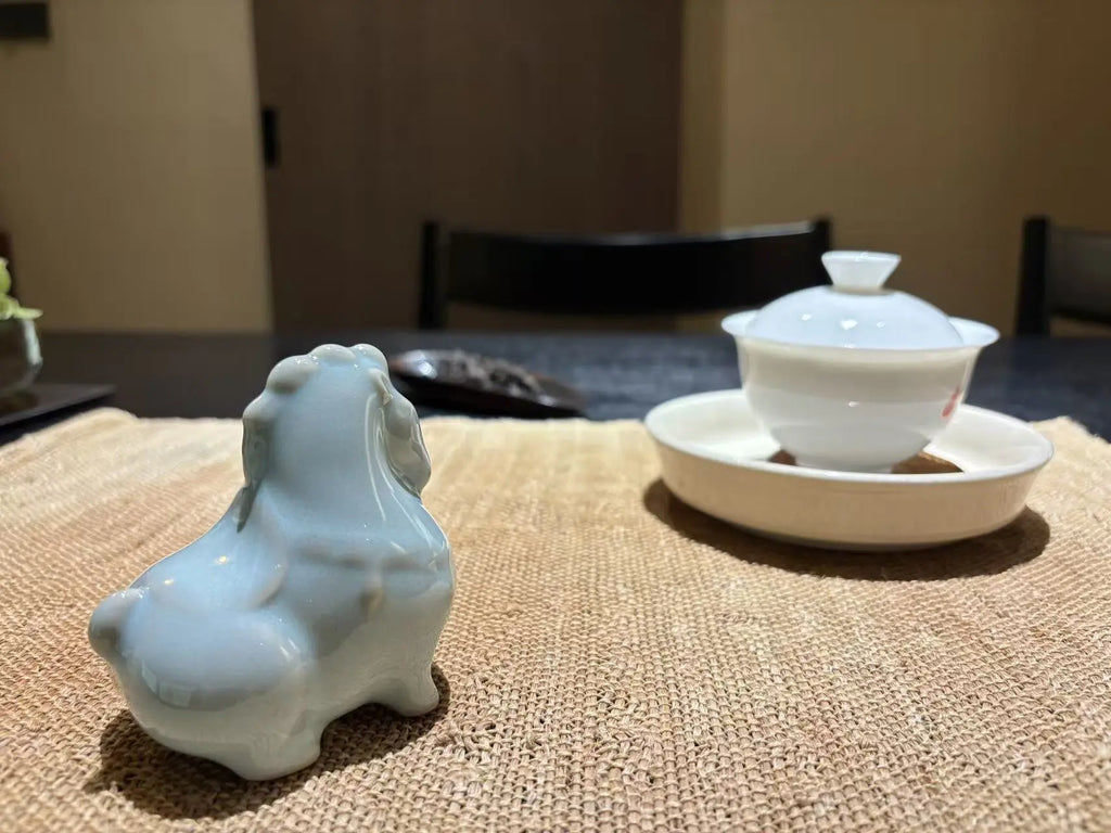 A small glossy pale blue ceramic horse tea pet in the foreground on a woven beige placemat, with a white lidded teacup and saucer blurred in the background on a dark table in an indoor setting.