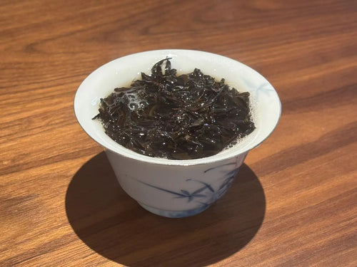 A white ceramic tea gaiwan with blue floral patterns, filled with Jin Jun Mei tea leaves, set on a wooden table.