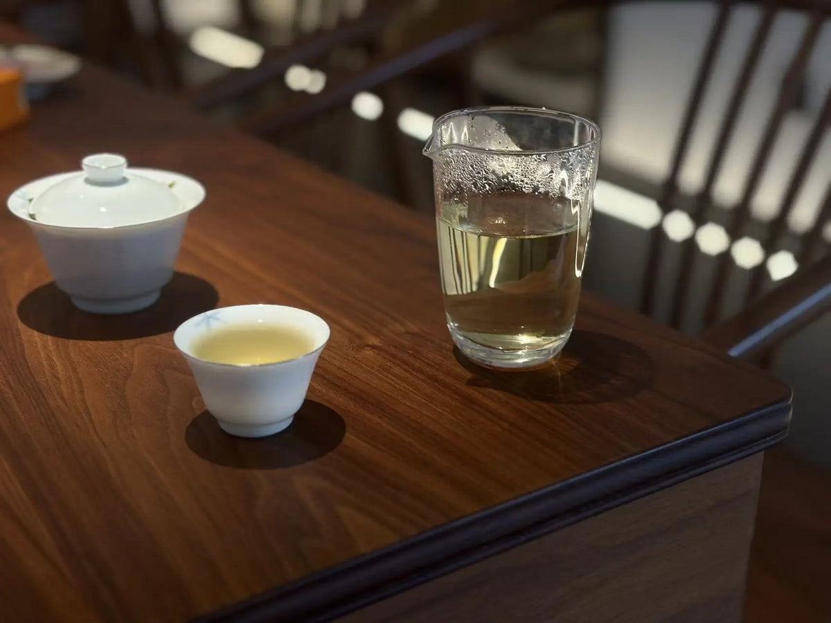 A glass tea infuser pitcher filled with pale tea, alongside a small white teacup and a white lidded teapot, arranged on a polished dark wood table in a softly lit interior setting.