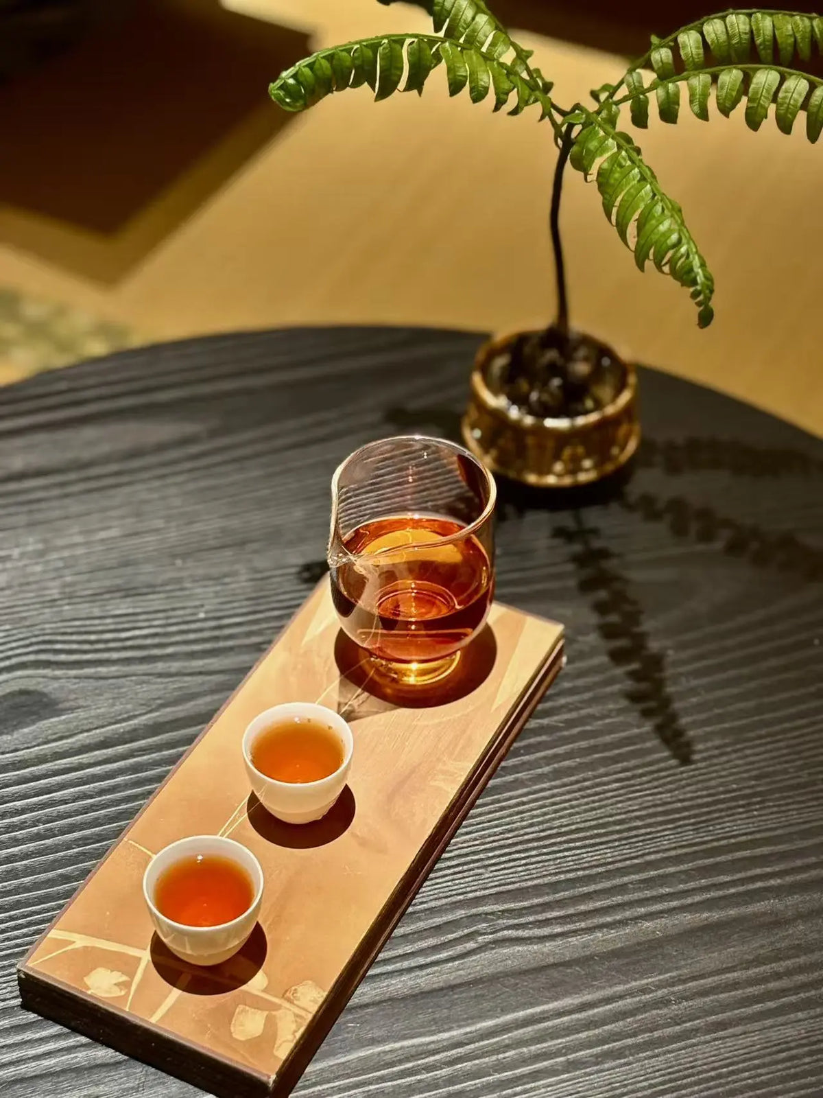 A glass pour-over tea infuser and two small white teacups filled with amber lapsang souchong tea liquor, arranged on a rectangular wooden tray on a dark round table. A potted fern sits in the background.