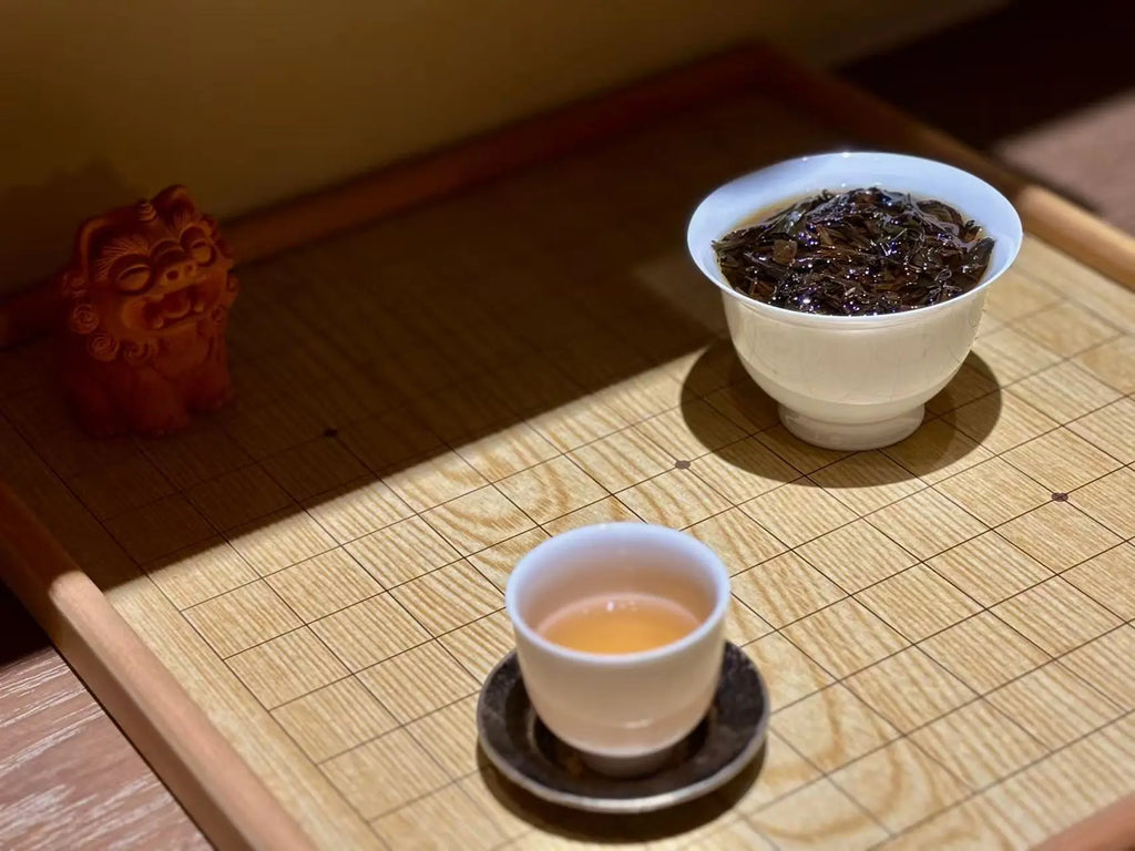 A traditional tea setup on a bamboo tray, featuring a white gaiwan filled with dark tea leaves and a smaller white teacup filled with amber Narcissus tea liquor, alongside a small red decorative figurine.
