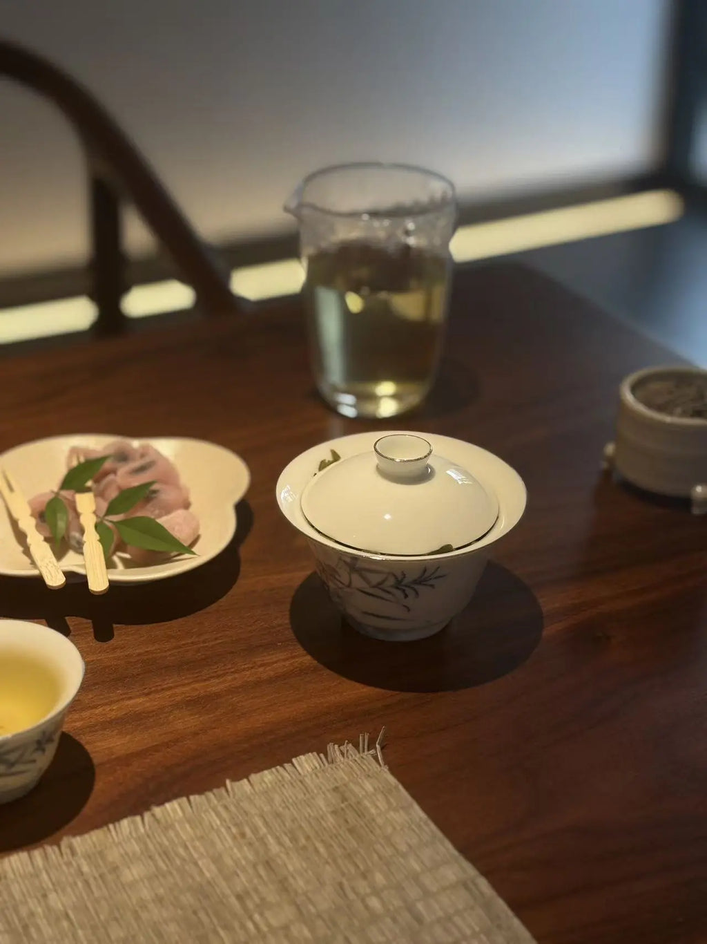 A white ceramic lidded tea bowl filled with iron goddess of mercy Oolong tea liquor with blue floral patterns sits on a dark wooden table, surrounded by tea-related items including a glass pitcher, a small plate with green leaves and pink slices, and a woven placemat.