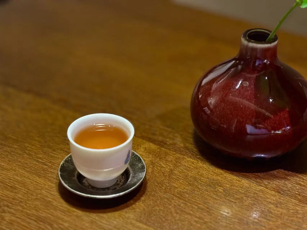 A small white teacup filled with amber Narcissus tea liquor sits on a dark saucer on a wooden table, next to a glossy dark red ceramic vase with a green stem.