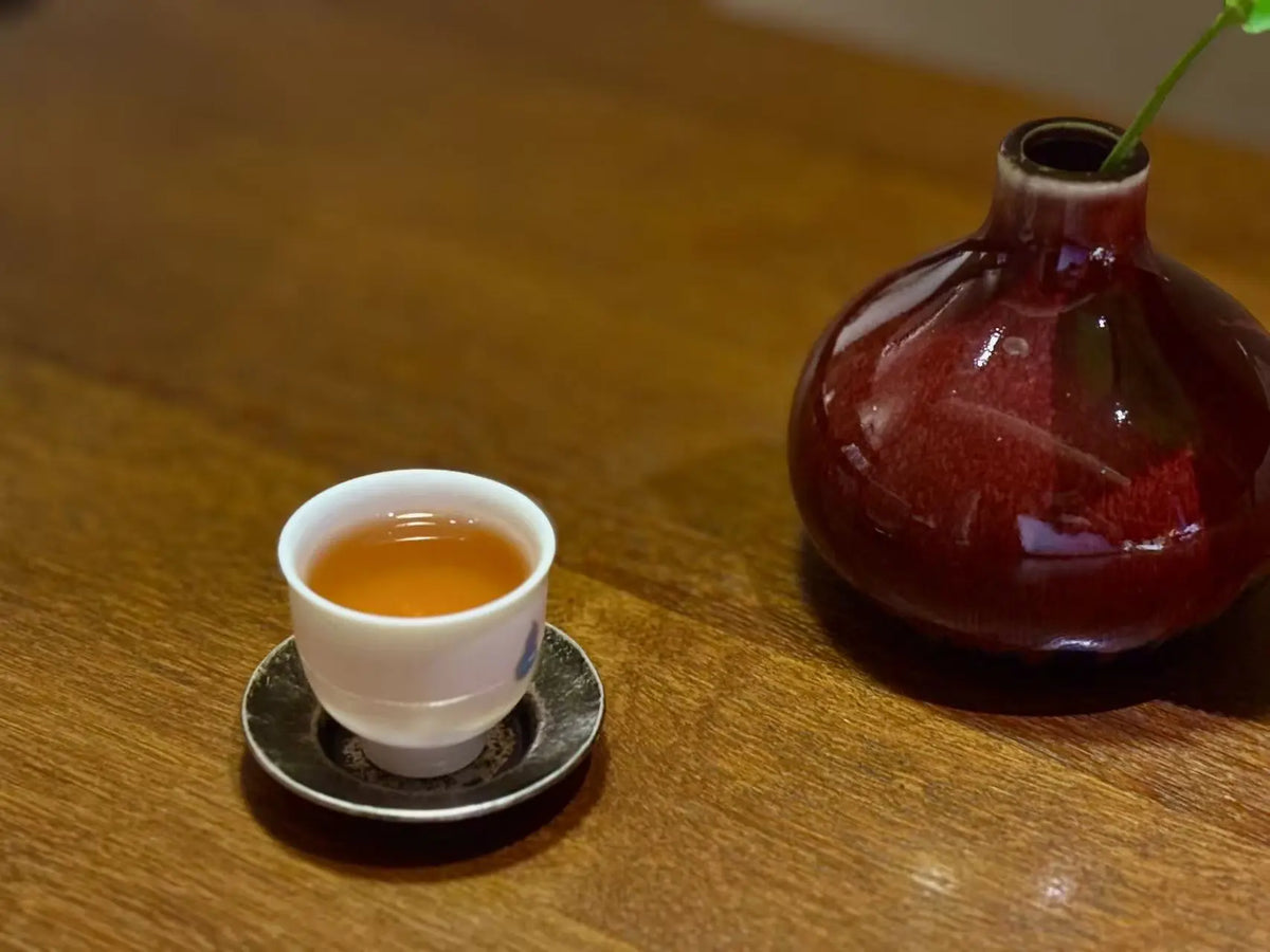 A small white teacup filled with amber Narcissus tea liquor sits on a dark saucer on a wooden table, next to a glossy dark red ceramic vase with a green stem.