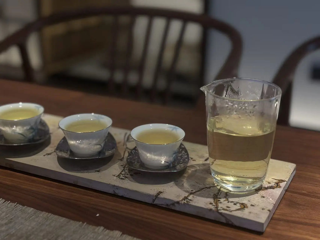 A glass pitcher filled with pale yellow tea sits on a marble tray beside three small white teacups with blue floral detailing on saucers, arranged on a wooden table with blurred chairs in the background.