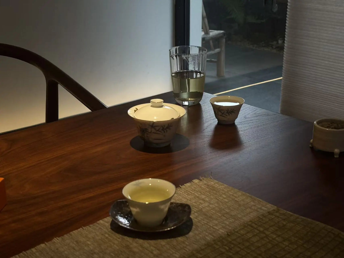 A traditional Chinese tea set arranged on a dark wooden table, featuring a lidded teapot, a small bowl, and two teacups with tea, alongside a glass of water, in a dimly lit indoor setting.