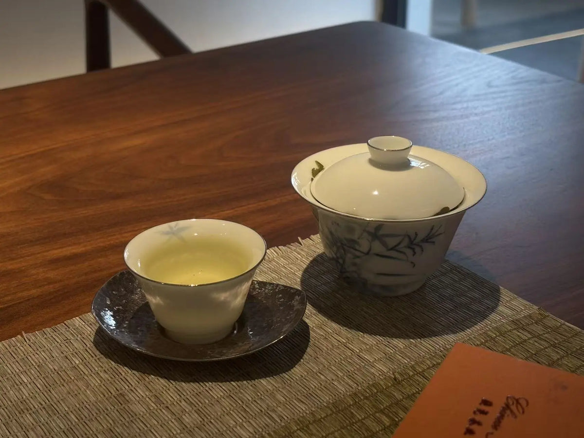 A ceramic tea set with a lidded gaiwan and a small cup on a saucer, filled with iron goddess of mercy tea placed on a woven placemat on a dark wooden table.