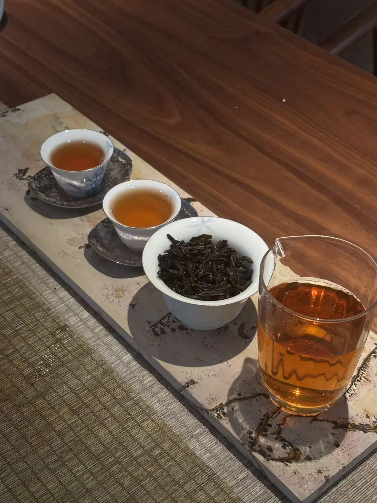 A Jin Jun Mei Tea serving setup on a wooden table, featuring two small white cups filled with amber tea, a white bowl of loose dark tea leaves, and a clear glass pitcher containing tea, arranged on a rectangular tray.