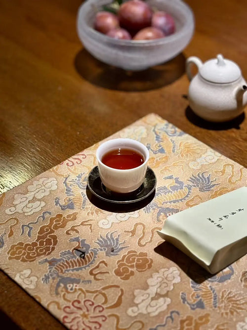 A small white teacup filled with amber Cinnamon rock tea liquor sits on a black saucer atop an ornate beige patterned fabric mat. A folded white ceramic tea towel or napkin lies beside it on the mat, with a white teapot and a bowl of red fruits blurred in the background on a wooden table.