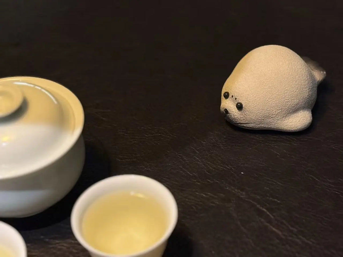 A small beige seal tea pet with black eyes sits on a dark wooden surface next to white ceramic tea cups and a teapot.