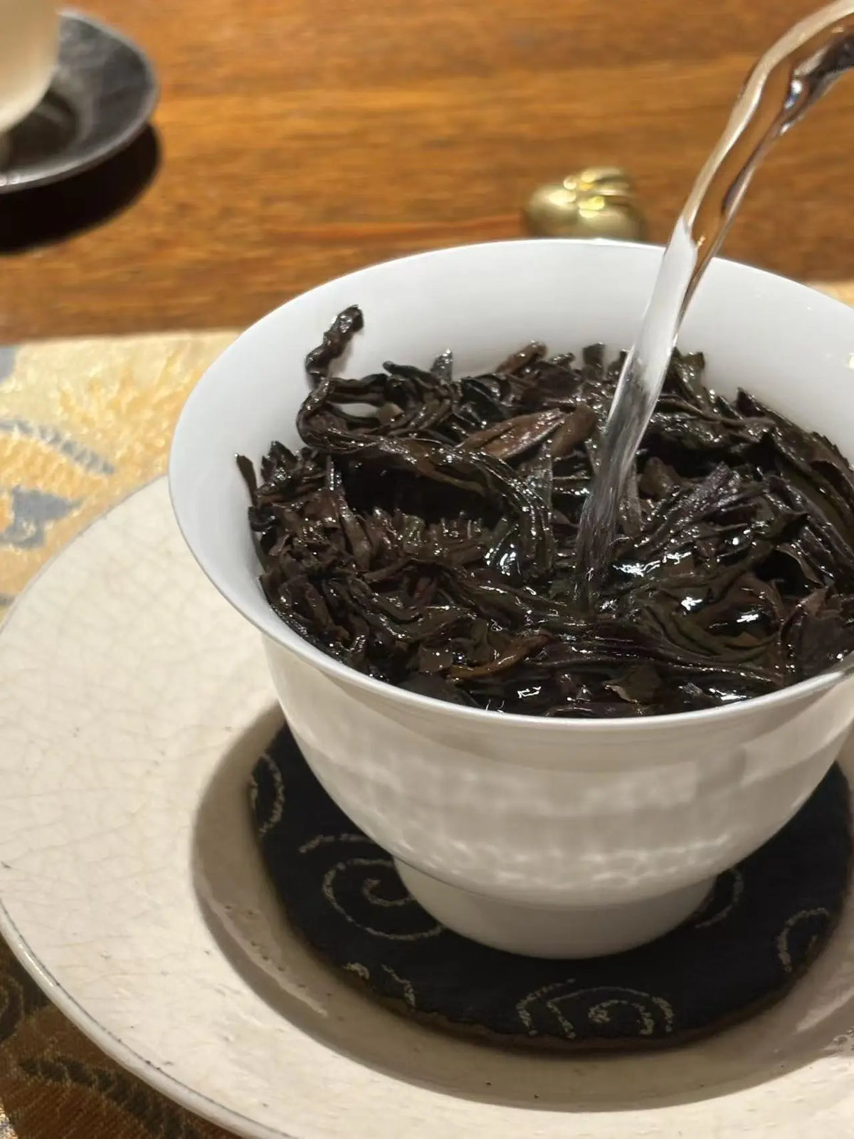 A white ceramic teacup filled with Da Hong Pao(Big Red Robe) tea leaves, with hot water being poured into it, set on a patterned coaster over a wooden table.