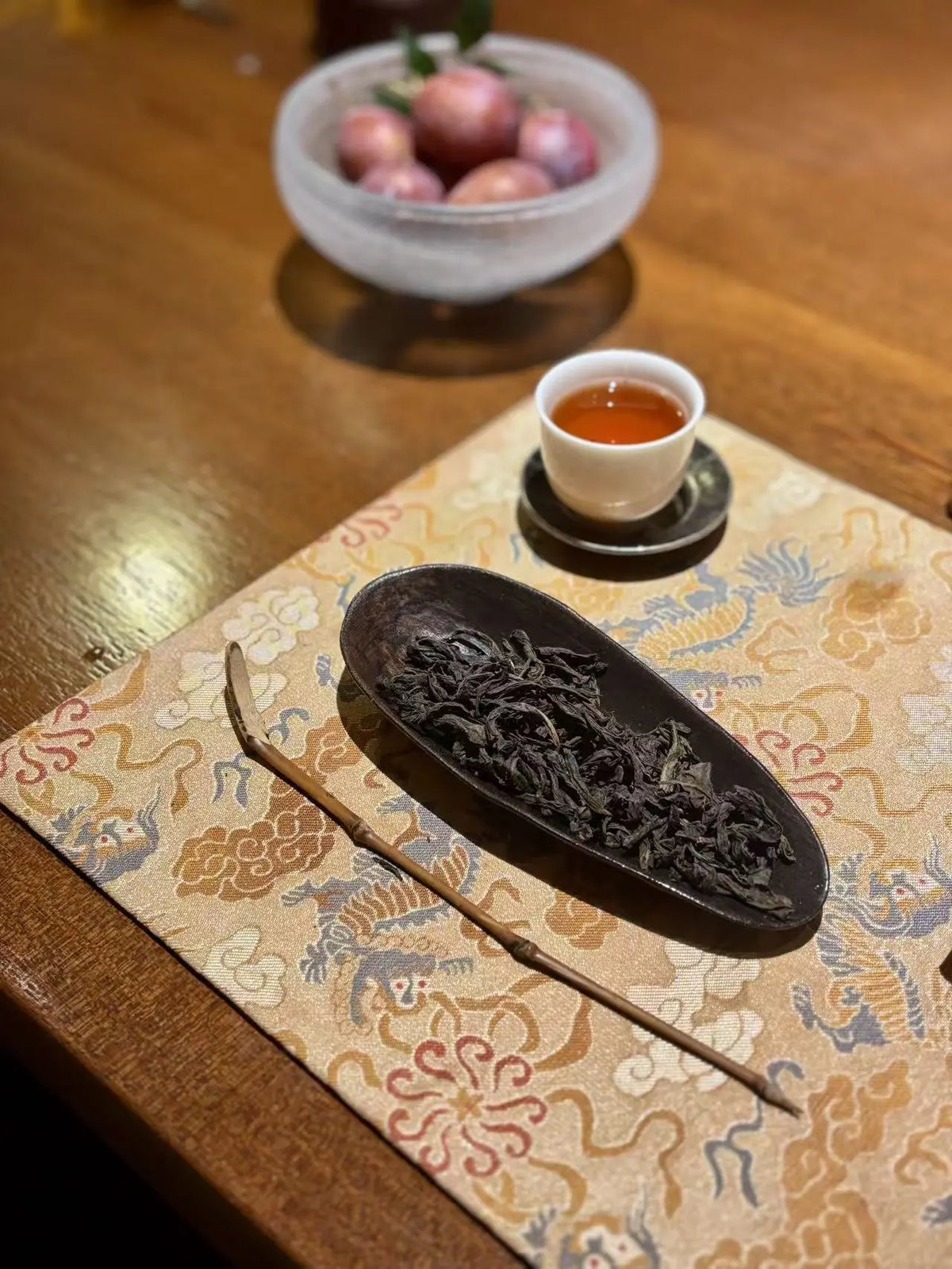 A close-up of lapsang souchong loose-leaf tea leaves in a dark oval dish, placed on a patterned beige fabric mat on a wooden table, with a small cup of amber tea and a bowl of red apples in the background.