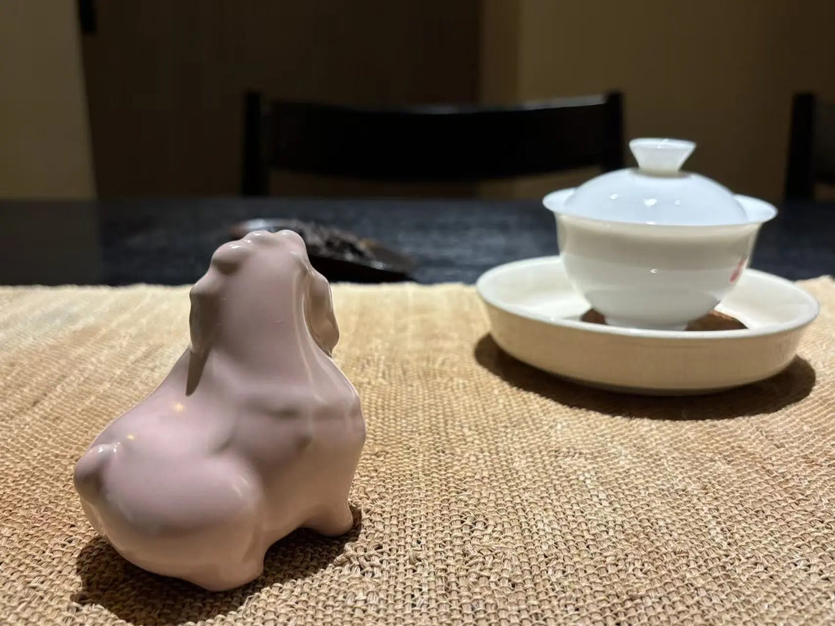 A small glossy pale pink ceramic horse tea pet, placed on a woven beige mat beside a white lidded teacup and saucer on a dark table.