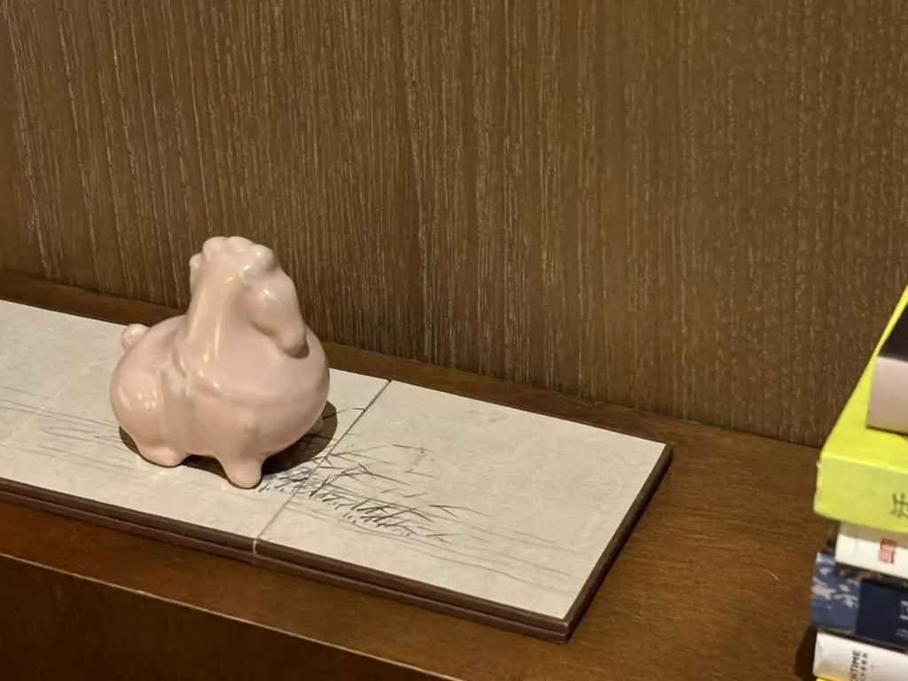 A small glossy pink ceramic horse tea pet, displayed on a rectangular light stone plaque on a wooden shelf against a brown textured wall.