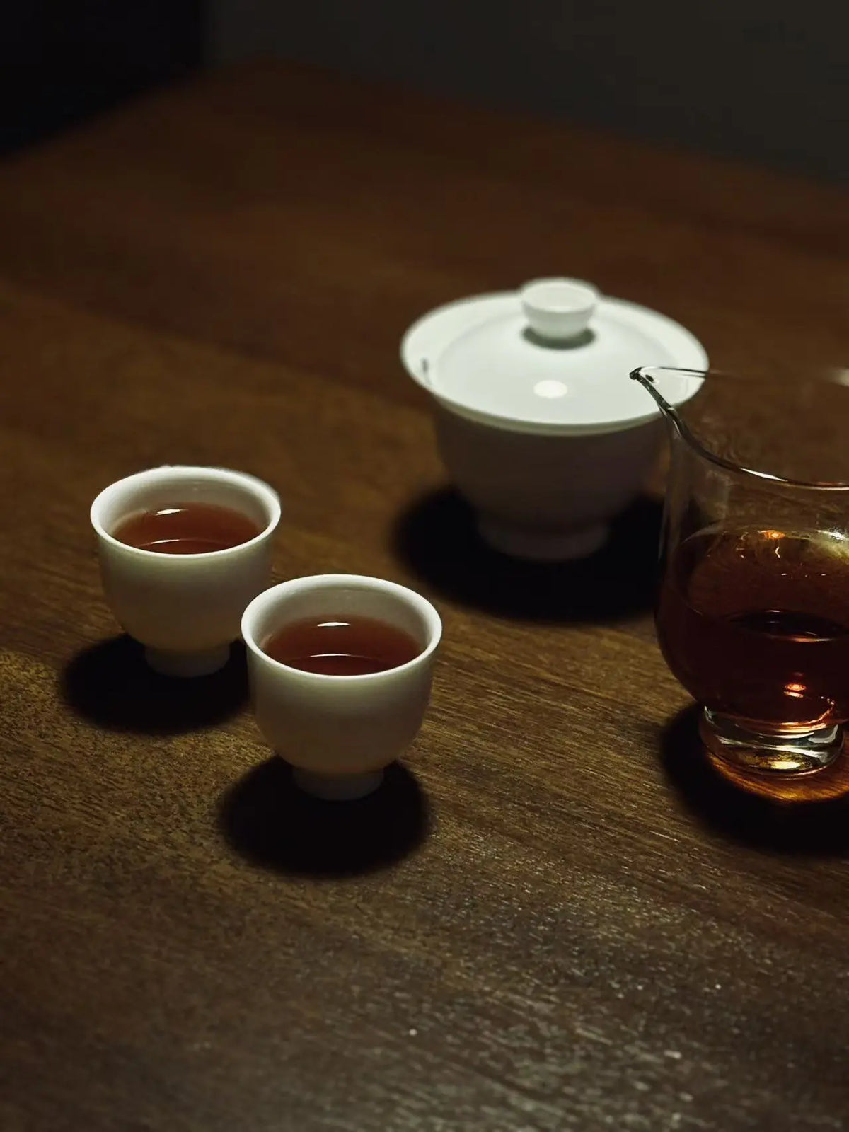 A white ceramic tea set on a dark wooden table, featuring two small cups filled with amber Narcissus tea liquor, a white lidded teapot, and a glass pitcher with tea, lit by warm directional light that casts strong shadows.