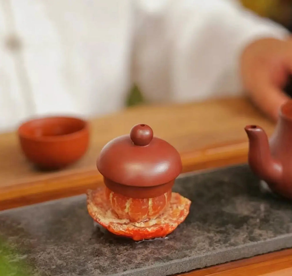 A close-up of a traditional Chinese clay teapot with a lid, resting on a orange tea pet and stone tray on a wooden table, with a blurred background showing a person in white clothing and another cup.