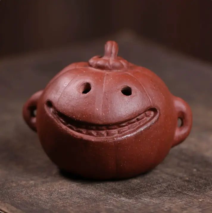 A small reddish-brown ceramic pumpkin-shaped vessel with a carved smiling face, photographed close up on a dark textured surface.
