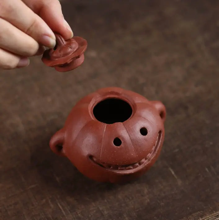 A close-up of a small reddish-brown ceramic teapot with a smiling face and side handles, shown on a dark wooden surface. A hand is lifting the lid off the teapot.