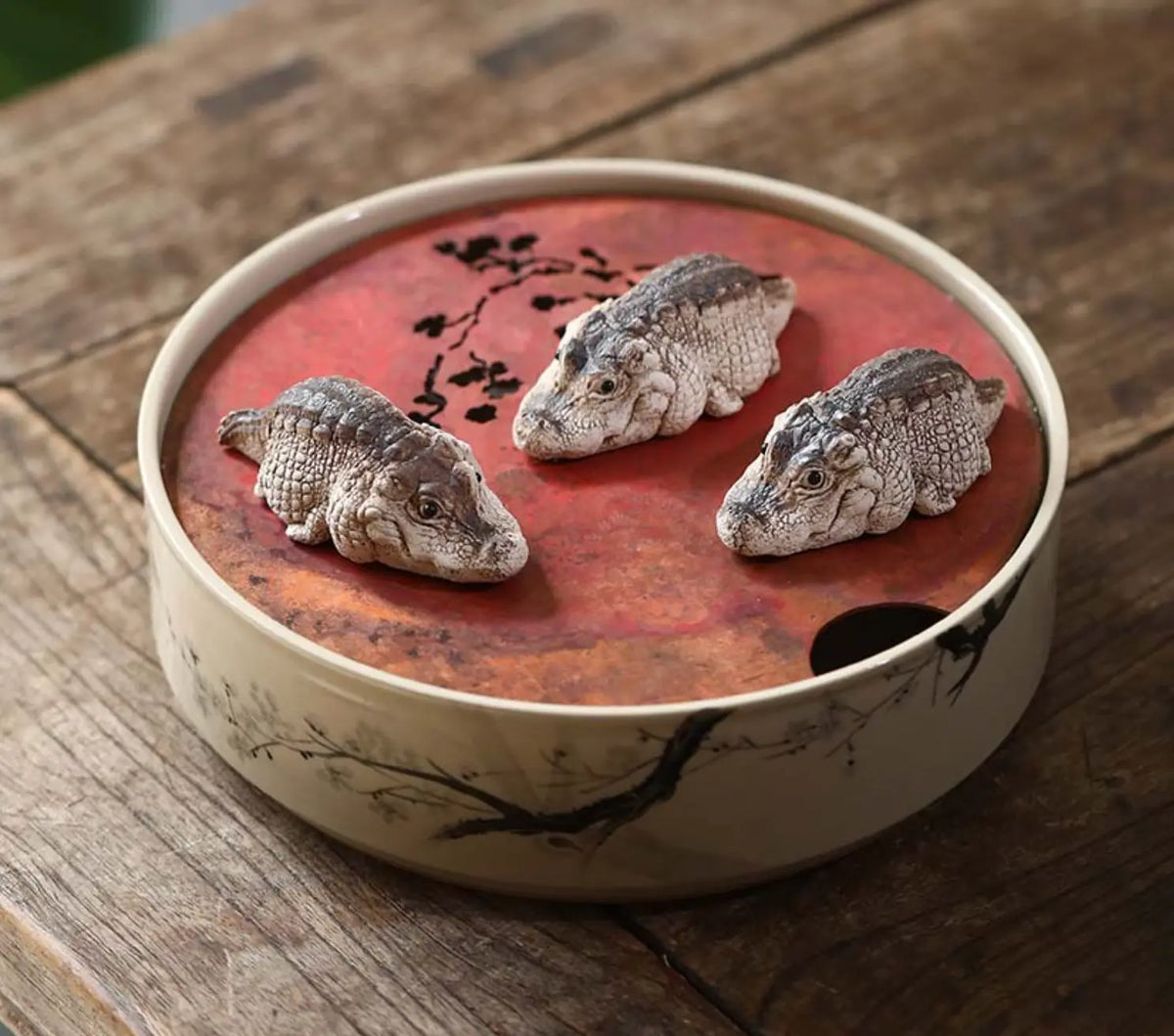 A round ceramic incense burner with a red lid featuring three small alligator tea pet set on a rustic wooden table.