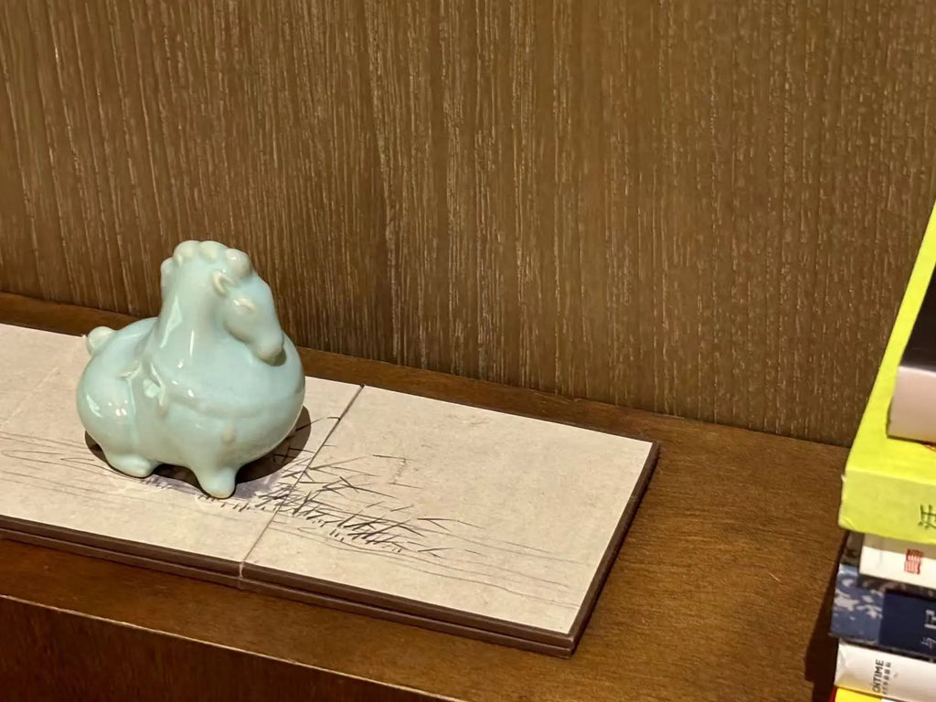 A small glossy light blue ceramic horse tea pet displayed on a rectangular stone tile coaster on a wooden shelf, with a textured brown wood-paneled wall behind it.