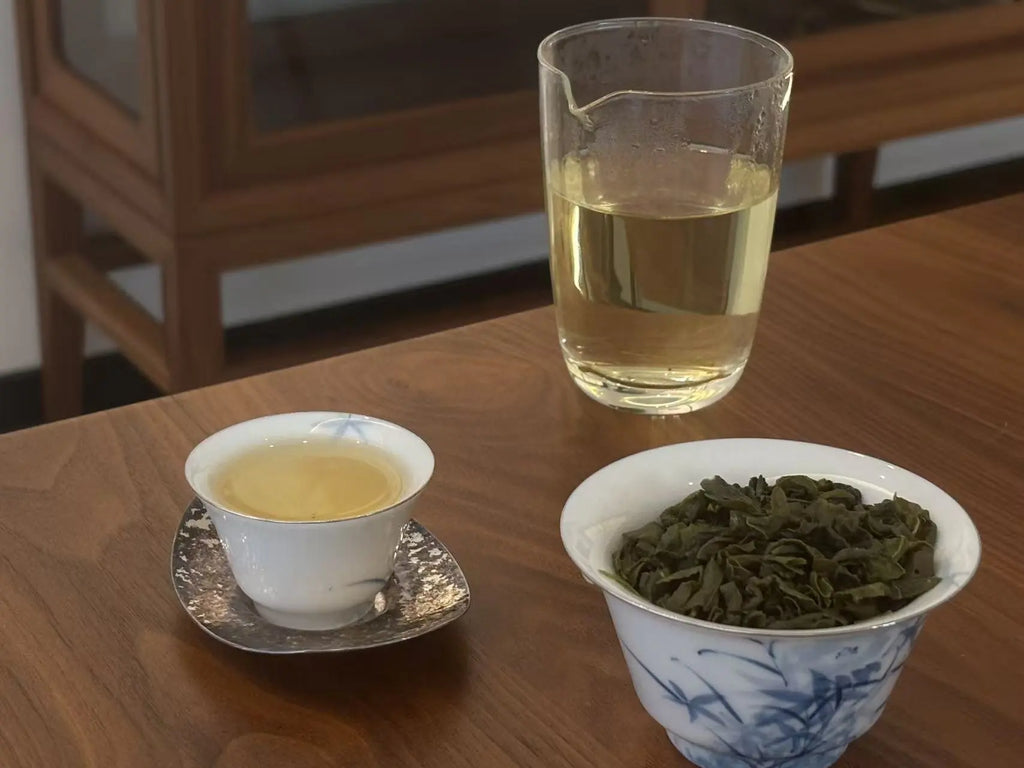 A glass pitcher filled with pale yellow tea, a small white teacup on a saucer with tea inside, and a blue-and-white bowl filled with dried green tea leaves, arranged on a wooden table.