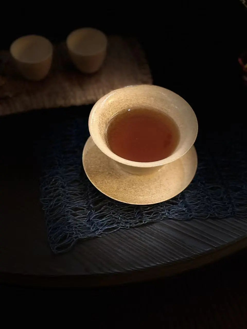A ceramic tea bowl and saucer set with amber Da Hong Pao (Big Red Robe) tea inside, styled on a dark textured mat with two small cups blurred in the background on a dark surface.