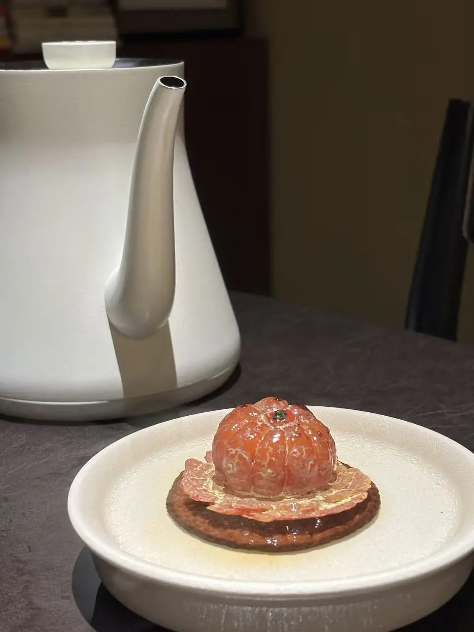 A close-up of a resin orange tea pet presented on a small plate in a real indoor setting, with a white kettle in the background on a dark tabletop.