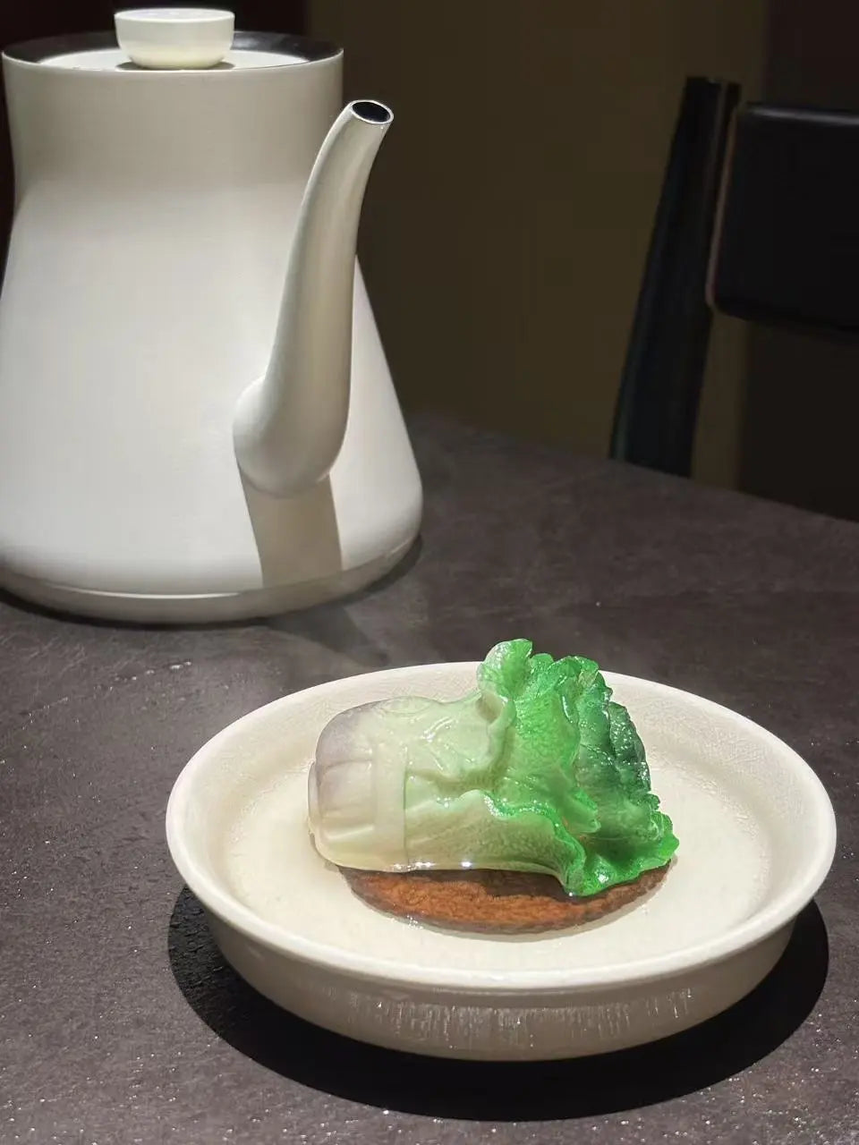 A carved green and pale cabbage  tea pet resting on a small round dish, with a white teapot blurred in the background on a dark tabletop.
