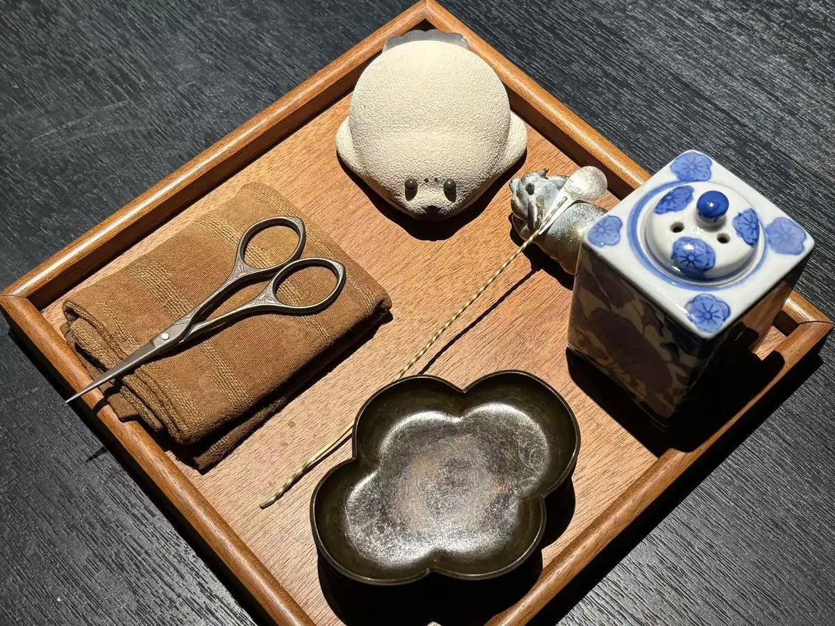 A styled flat-lay of traditional Japanese tea ceremony tools arranged on a wooden tray, including a small ceramic pitcher, metal scissors on a folded cloth, a flower-shaped dish, and a ceramic seal tea pet, set against a dark wood background.