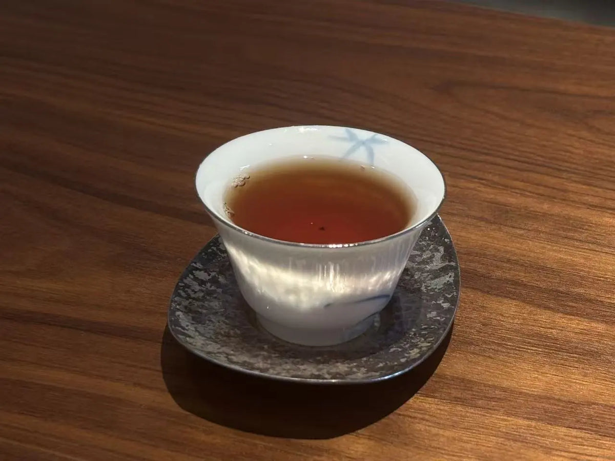 A white ceramic tea cup filled with amber-red Lapsang souchong tea liquor, resting on a dark speckled saucer on a wooden table.