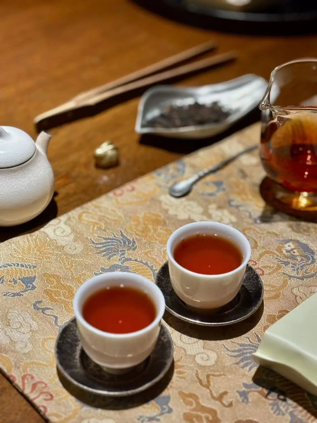 Two white teacups filled with amber Da Hong Pao(Big Red Robe) tea liquor, set on a patterned textile over a wooden table with a teapot, chopsticks, and a glass pitcher nearby.