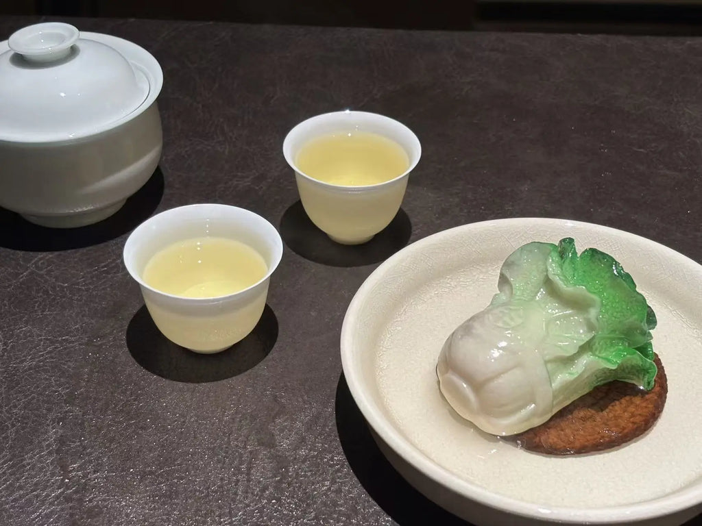 A ceramic tea set with a white lidded teapot and two small cups filled with pale yellow tea, arranged on a dark tabletop beside a plate holding a cabbage tea pet on a brown base.