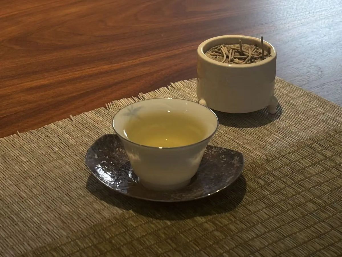 A white ceramic tea cup filled with pale yellow Iron Goddess of Mercy tea sits on a dark saucer, placed on a woven placemat over a wooden table. A small white pot containing dried tea leaves is behind the cup.